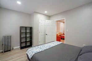 Quarto com cama, radiador, guarda-roupa e porta aberta para outro espaço.