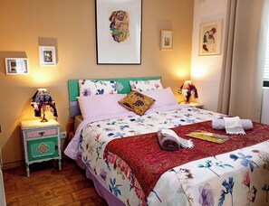 Een netjes opgemaakt bed met bloemenbeddengoed, een nachtkastje in het groen met een lamp en een ingelijst plaatje aan de muur.