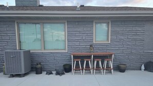 A patio area with a stone wall, two windows, a table with four stools, and outdoor air conditioning units. - 'Honey Bee' Family Home in Columbus, Nebraska! (Columbus)