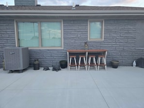 Un patio con pared de piedra, dos ventanas, una mesa y cuatro taburetes, además de unidades de aire acondicionado exteriores.