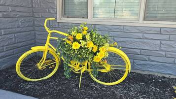 Uma bicicleta amarela com arranjo floral, posicionada em frente a um edifício com janela.