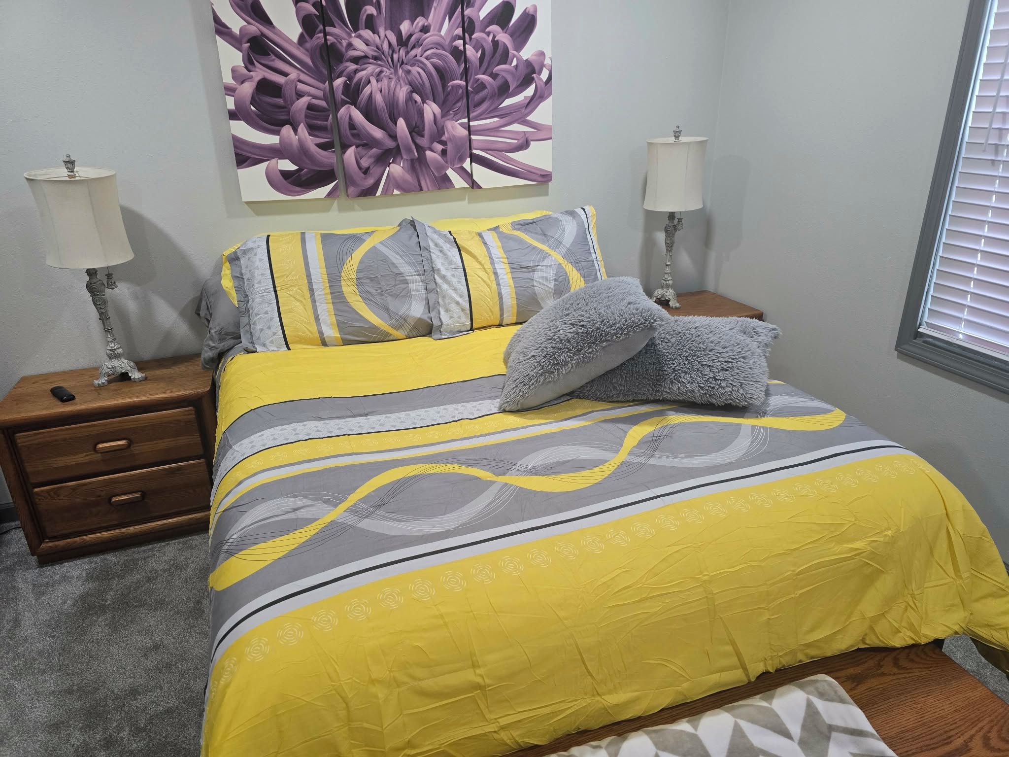 Quarto com cama, duas mesas de cabeceira, abajur, grande pintura floral na parede e janela com persianas.