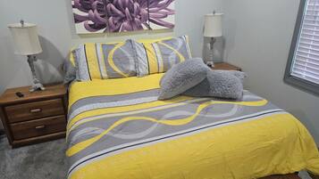 Quarto com cama, duas mesas de cabeceira, abajur, grande pintura floral na parede e janela com persianas.