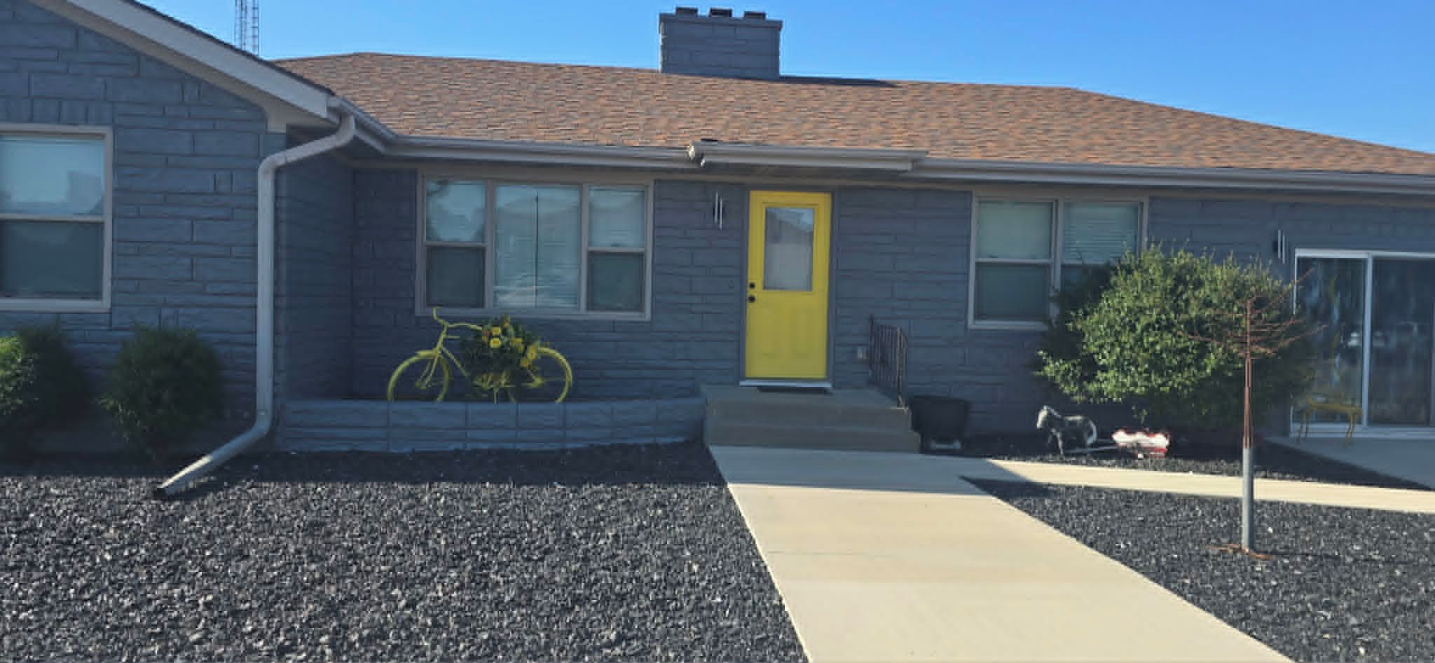 Une maison d’habitation avec une porte jaune, une cour avant recouverte de gravier, un petit arbre et un vélo.