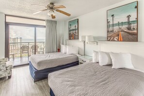 Habitación de hotel con dos camas, un balcón con sillas, ventilador de techo y dos cuadros con motivos playeros.