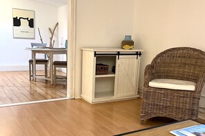 A room with a wicker chair, a white cabinet with a sliding door, a dining table with chairs, and a framed picture on the wall. - Alter Konsumverein, App No. 1 - Your Comfortable Apartment at Alter Konsumverein (Sylt)