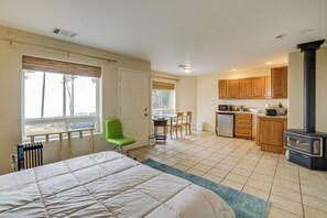 A bedroom with a bed, a green chair, a dining area with a table and chairs, a kitchen with wooden cabinets, and a wood-burning stove.