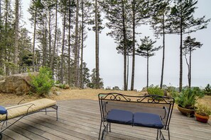 A wooden deck with two metal chairs and a bench, overlooking a forest and a body of water.