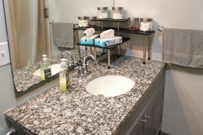 Casa de banho com bancada de granito, lavatório branco, espelho, porta-toalhas e prateleira com artigos de higiene pessoal.