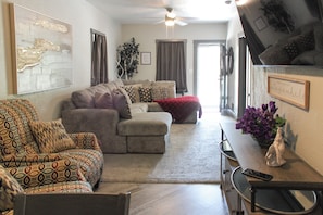Uma sala de estar com sofá, poltrona estampada, mesa de jantar e um vaso de flores, e ventilador de teto.