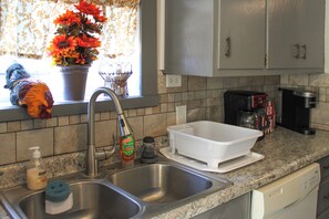A kitchen with a double sink, a coffee maker, a dish rack, and a window with a flower arrangement.
