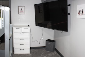 A room with a flat-screen TV mounted on the wall, a white dresser with drawers, and a small gray speaker on the floor.