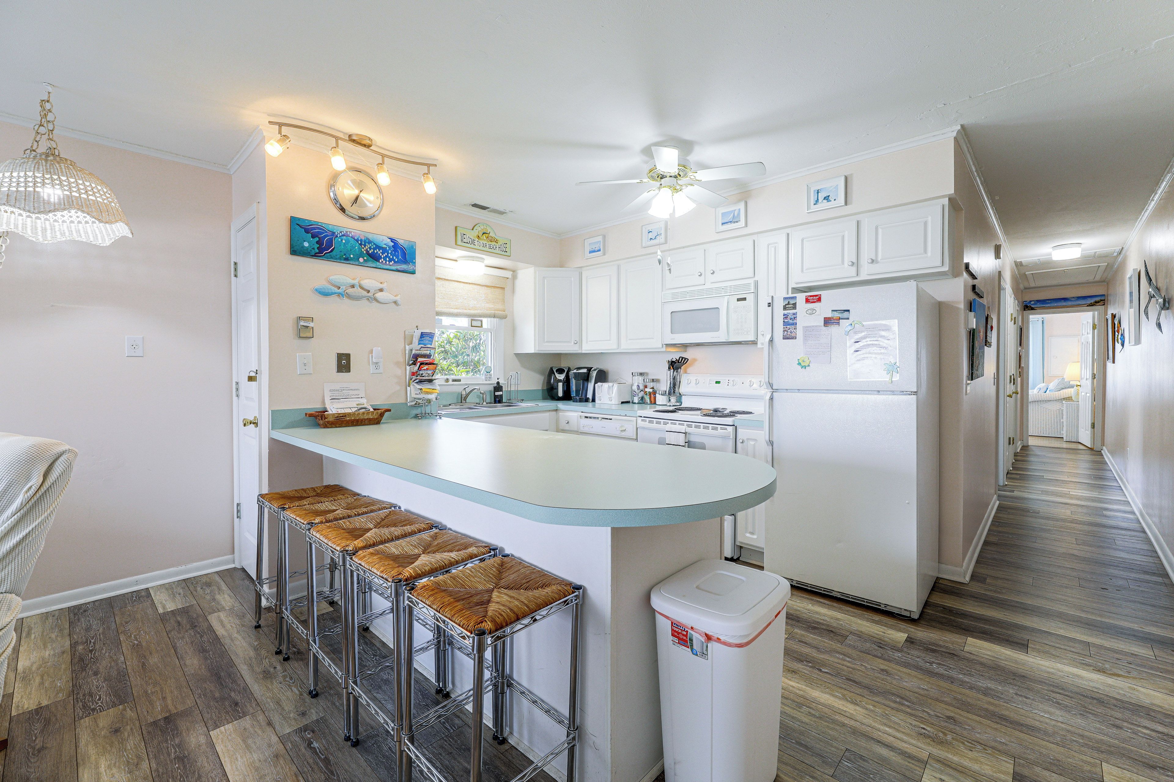 Una cocina moderna con una isla central, taburetes de bar, un ventilador de techo y vista a la playa a través de la ventana.