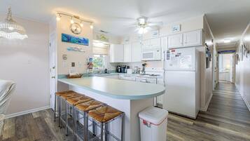 Una cocina moderna con una isla central, taburetes de bar, un ventilador de techo y vista a la playa a través de la ventana.