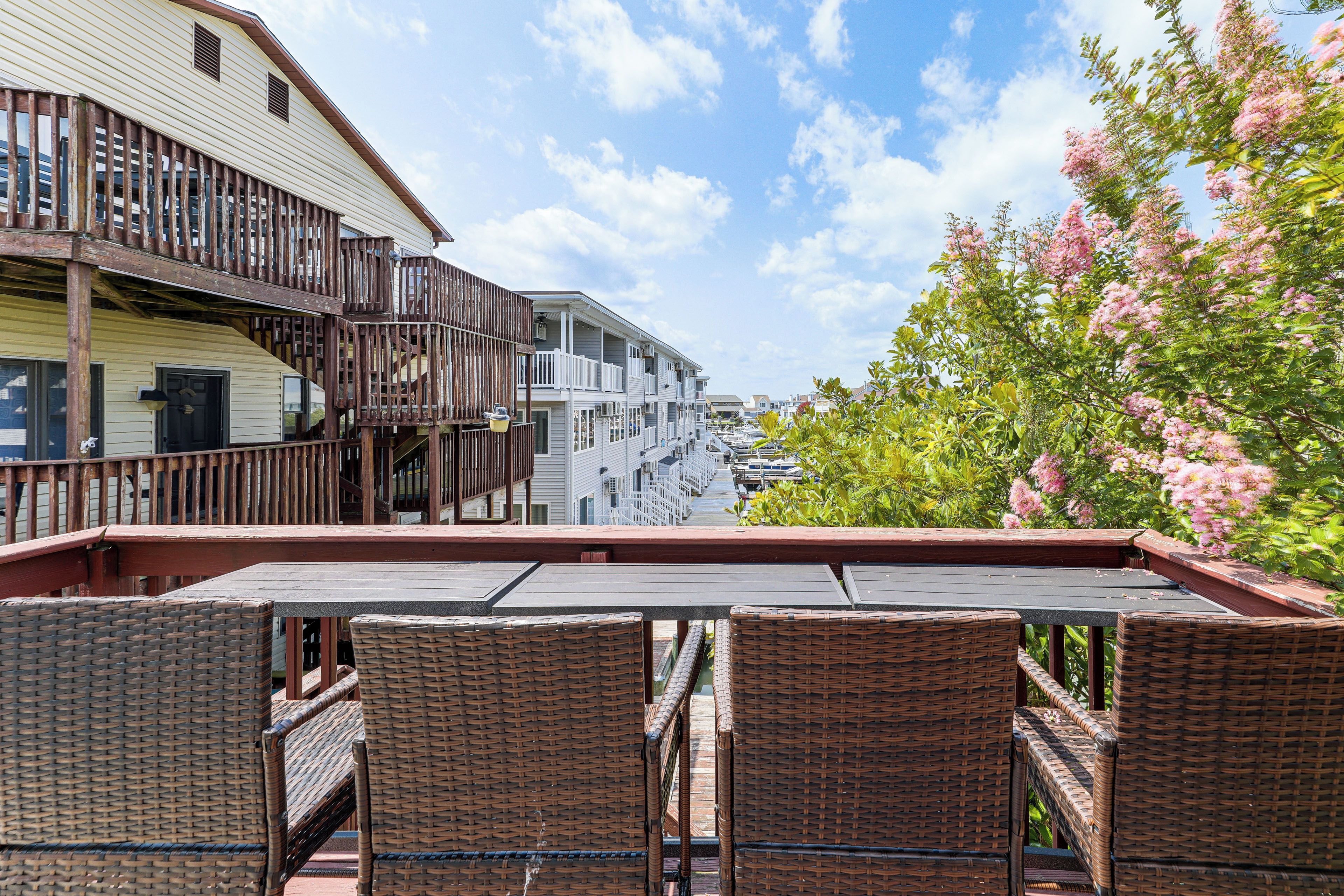 A balcony with wicker furniture overlooking a marina with boats, adjacent buildings, and blooming pink trees.