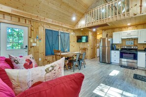 A cozy cabin interior with a dining area, kitchen, and a red couch with floral cushions.