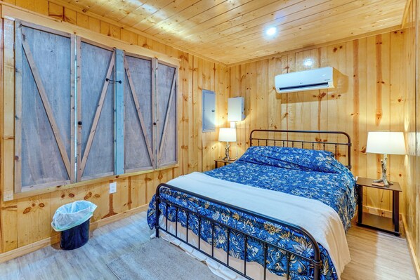 A wooden room with a bed, two bedside tables, a wall-mounted air conditioner, and a window with shutters.