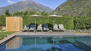 Piscina cristalina con sillones de descanso y sombrillas, rodeada de vegetación y montañas.