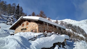 Un edificio de piedra con techo inclinado, rodeado de montañas y árboles cubiertos de nieve.