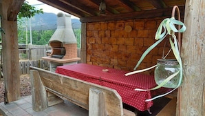 Un área de estar al aire libre con un horno de ladrillo, bancos de madera y una mesa cubierta con mantel rojo.