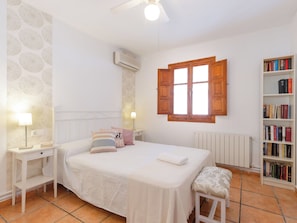 Une chambre à coucher comprenant un lit, des tables de chevet, une lampe, une fenêtre avec des volets en bois, un radiateur et une bibliothèque.