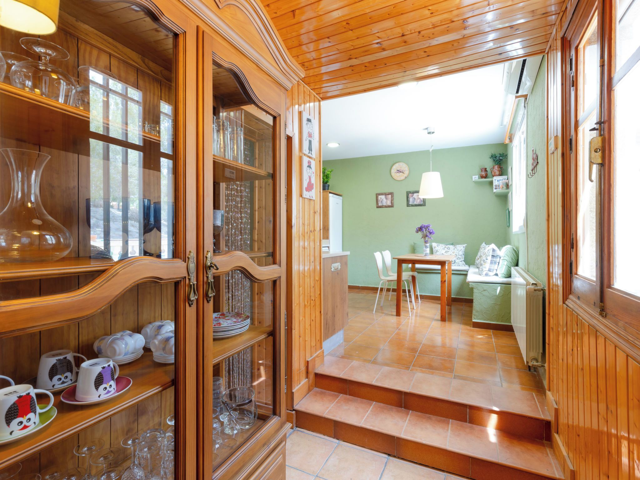 Un intérieur en bois, avec un meuble vitré rempli de verres, un coin repas avec une table et des chaises, et un escalier menant à une autre pièce.