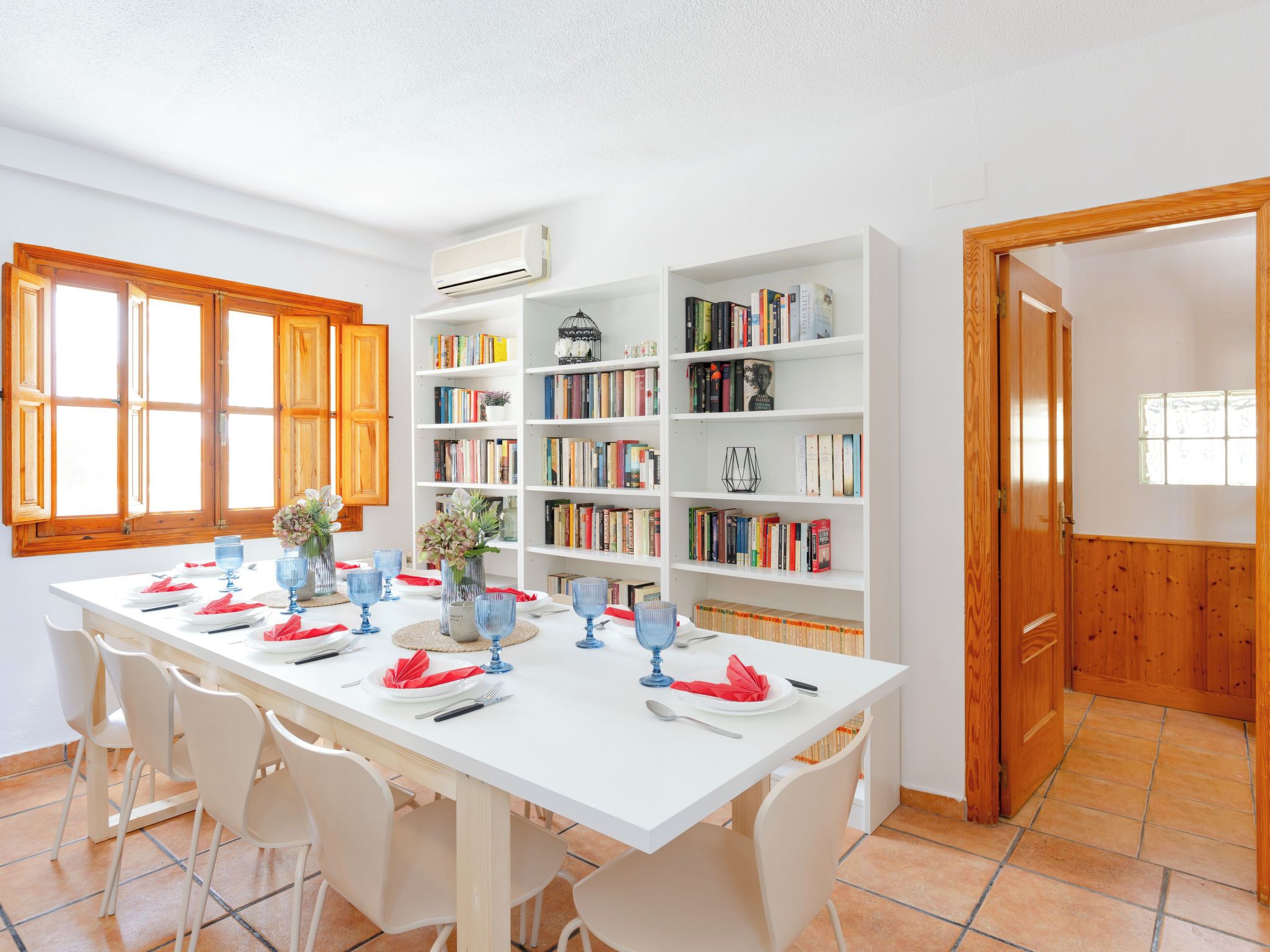 Un coin repas avec une table blanche dressée pour six personnes, entourée de chaises blanches, et une bibliothèque remplie de livres sur le côté droit.