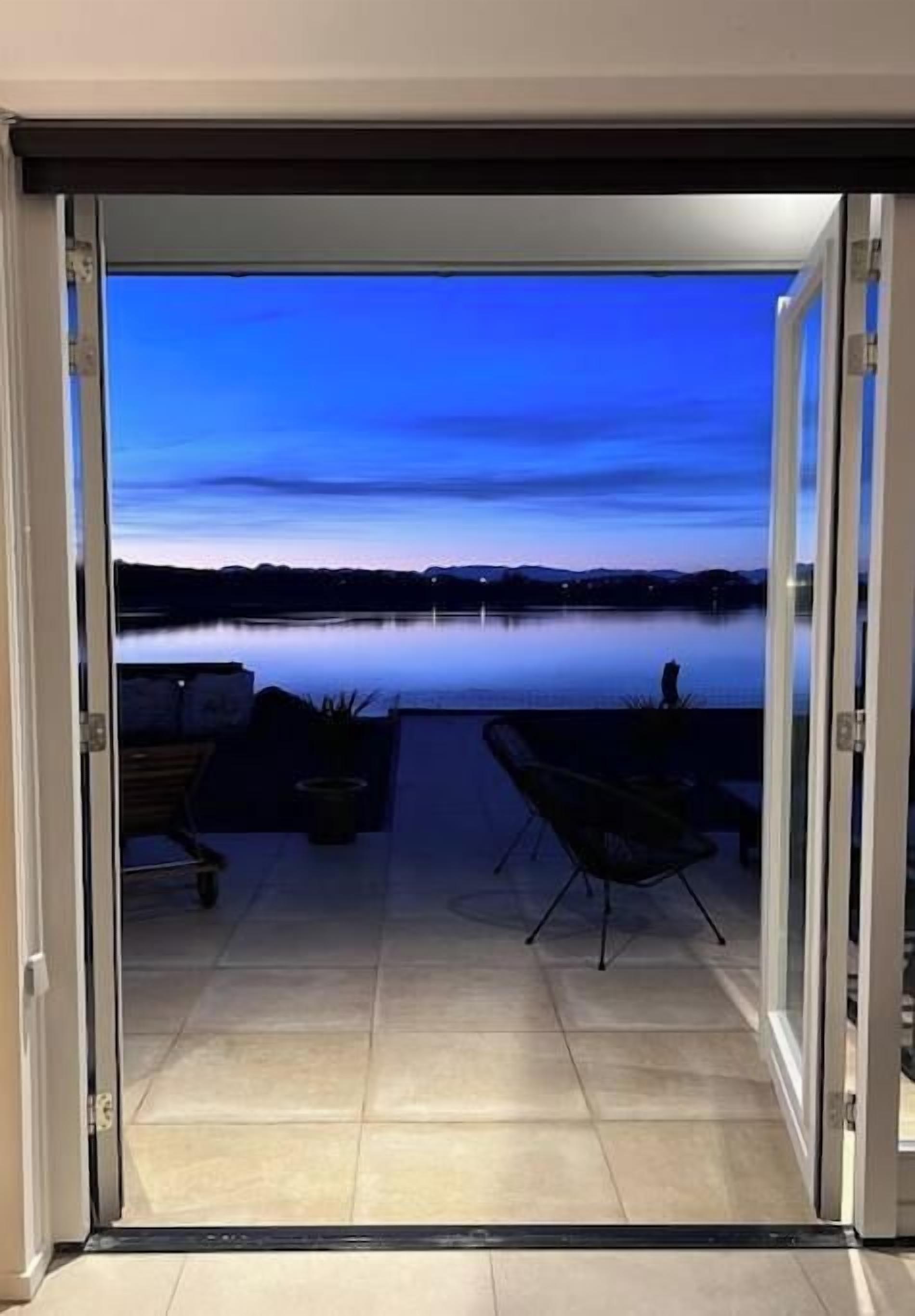 An open sliding door leading to a patio with a view of a lake at dusk.