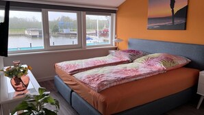 Ein Schlafzimmer mit einem Bett, einem Fenster mit Blick auf Boote, einer Pflanze und einem Gemälde.