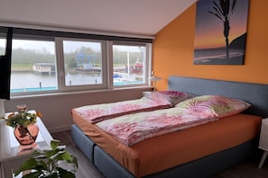 A bedroom with a bed, a window with a view of boats, a plant, and a painting. - Holiday home with 1 bedroom (Wieringerwerf)
