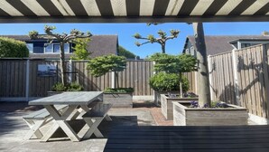 En overdækket terrasse med et træbord og bænke, omgivet af træhegn og krukker med planter.