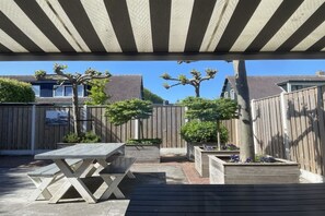 Een overdekt terras buiten, met een houten tafel en bankjes, omgeven door houten hekken en potplanten.