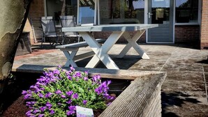 En patio med et træbord og stole, et blomsterbed med lilla blomster, og en bygning med store vinduer og en overdækket indgang.