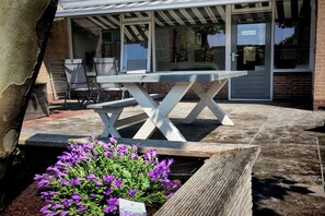 Een terras met een houten tafel en stoelen, een bloembed met paarse bloemen, en een gebouw met grote ramen en een overdekte ingang.