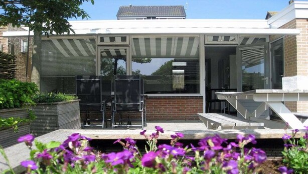 Et moderne hus med en patio, en glasdør der fører til en overdækket udendørs opholdsplads, og en murstensmur.
