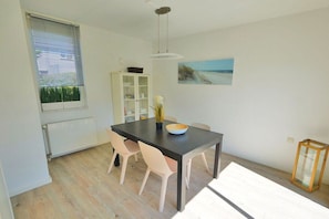 A dining area with a table, chairs, a cabinet, a painting, and a window with blinds. - De C"oogse Strandloper (Callantsoog)