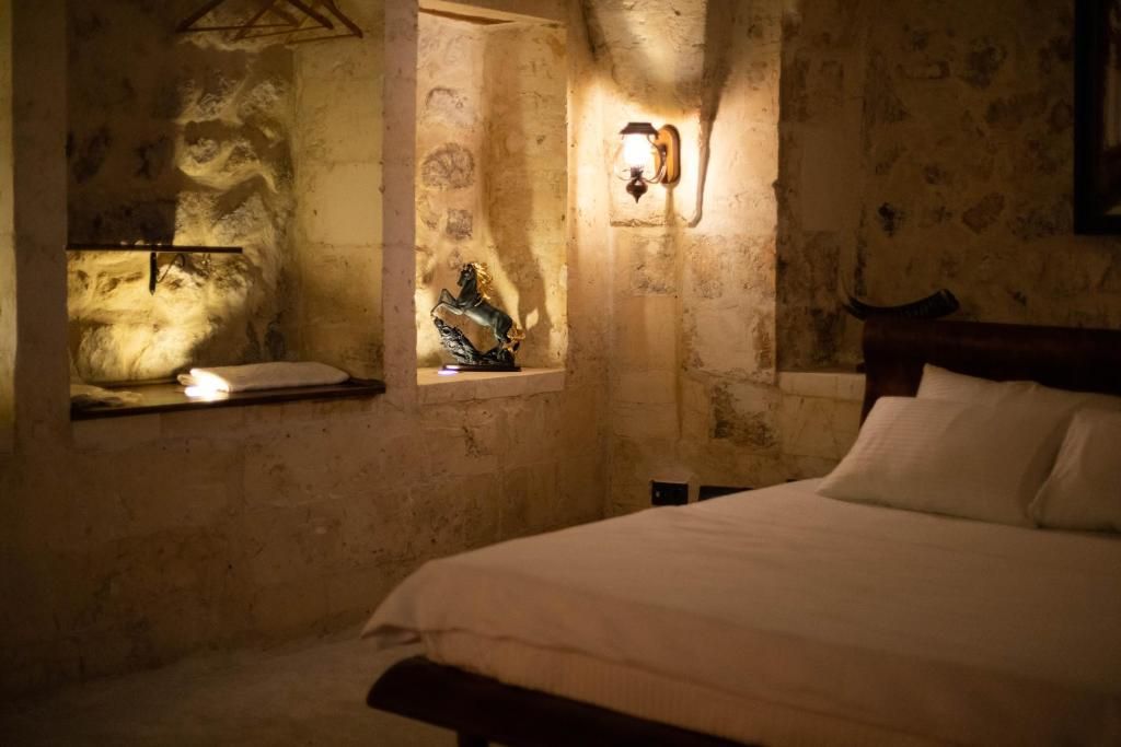 A bedroom with a stone wall, a bed with white bedding, a nightstand, a wall-mounted light fixture, and a window with a decorative item.
