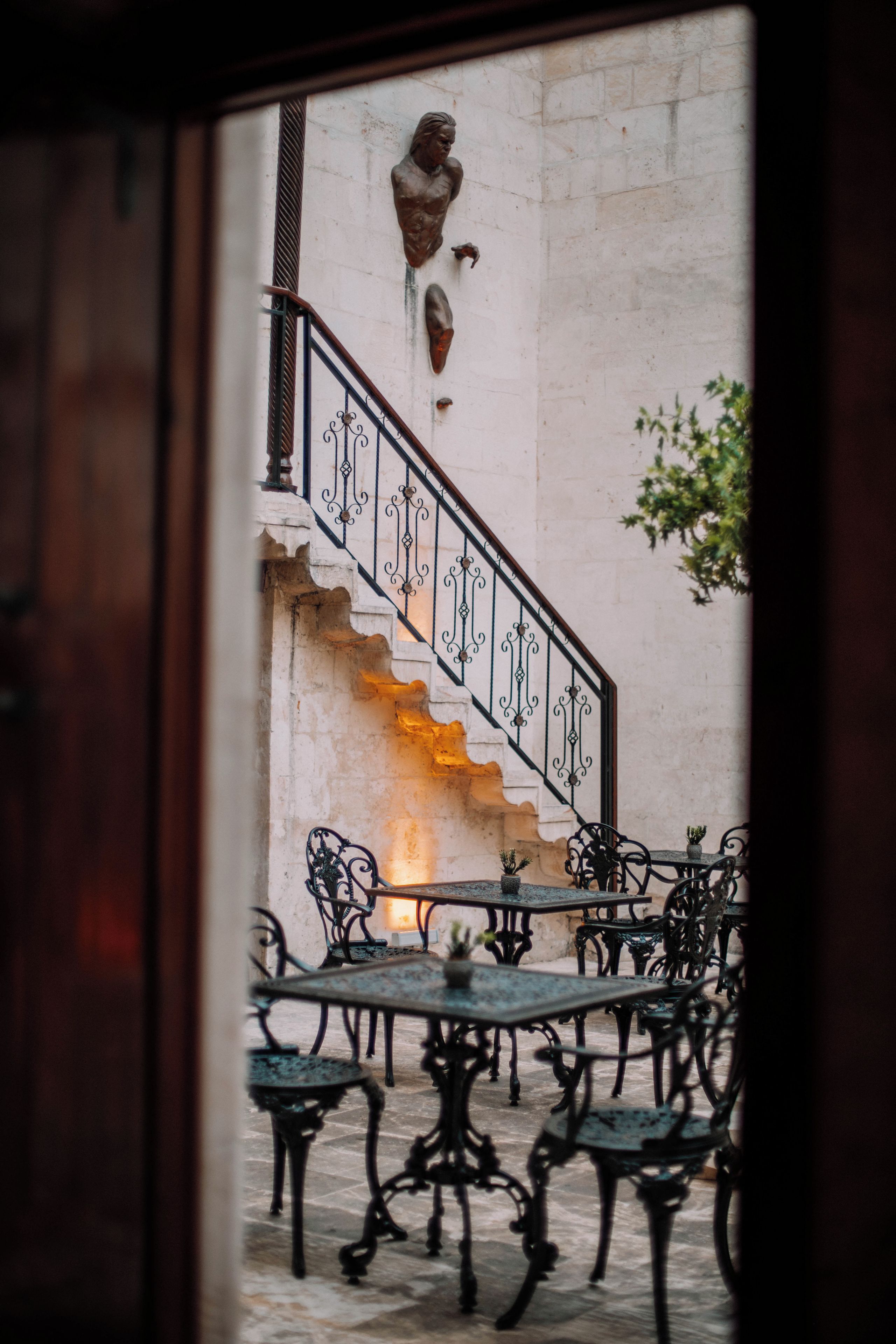 Una escalera con barandillas metálicas ornamentales que conduce a un nivel superior. Abajo, hay mesas y sillas al aire libre, lo que sugiere un área de patio o terraza.