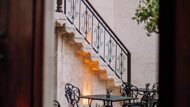 Una escalera con barandillas metálicas ornamentales que conduce a un nivel superior. Abajo, hay mesas y sillas al aire libre, lo que sugiere un área de patio o terraza.