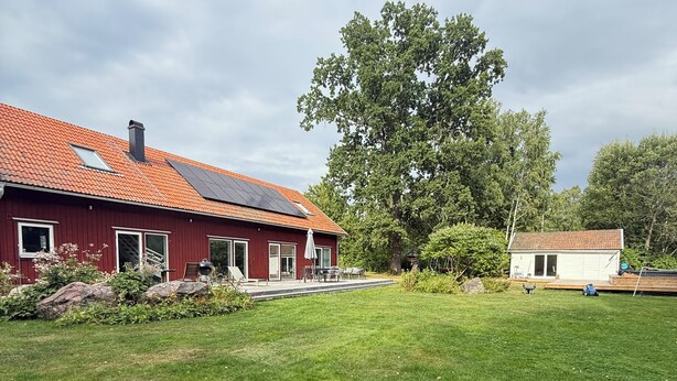 Una casa roja con un panel solar en el techo, un edificio blanco y un árbol grande en el patio.