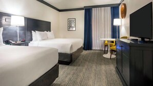 Ein Hotelzimmer mit zwei Betten, einem Schreibtisch mit Stuhl, einem Fernseher und einem Fenster mit Vorhängen.