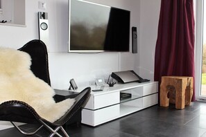 Ein modernes Wohnzimmer mit einem flachbildfernseher an der Wand, einem Glastisch, einem Ledersessel, einem Holzschemel und einem weißen Schrank.