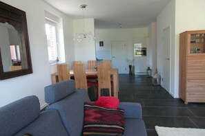 Ein Wohnzimmer mit einer Couch, einem Esstisch mit Stühlen, einer Küchenzeile im Hintergrund und einem großen Spiegel an der Wand.