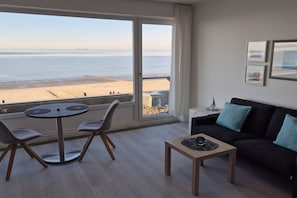A room with a large window overlooking a beach, a round table with chairs, a black sofa with blue cushions, and a small side table with a lamp. - Nautilus Penthouse Beach View (Cuxhaven)
