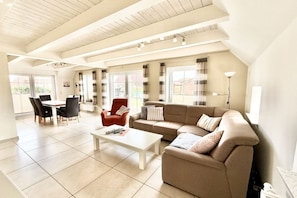 A spacious living area with a high ceiling, a sofa set, a dining table with chairs, and large windows with curtains. - Friesengeist Country House (Greetsiel)