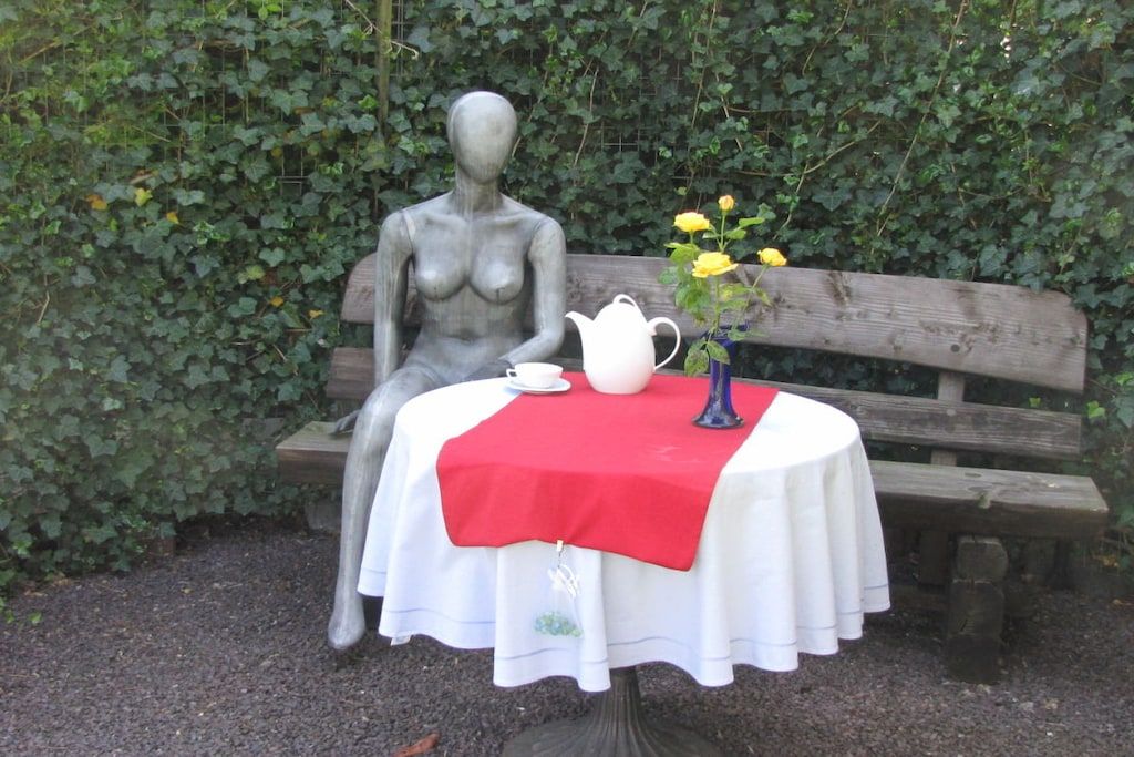 A statue of a woman seated on a bench with a table in front of it, covered with a white tablecloth and a red cloth, and a vase with yellow flowers.