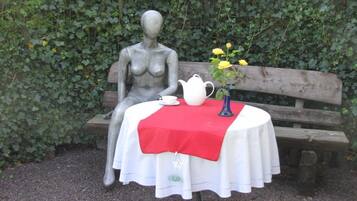A statue of a woman seated on a bench with a table in front of it, covered with a white tablecloth and a red cloth, and a vase with yellow flowers.