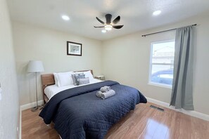 Quarto com cama, ventilador de teto, janela com cortinas e um quadro na parede.