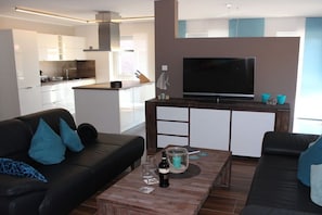 A modern living room with a black leather sofa, a coffee table with a bottle and glasses, and a TV on a wooden stand. - Holiday apartment with 1 bedroom (Krummhörn-Greetsiel)