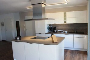 A modern kitchen with white cabinetry, a central island with a sink, and a stainless steel range hood. - Holiday apartment with 1 bedroom (Krummhörn-Greetsiel)